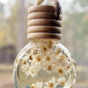Puede incluir: Un ambientador de coche de cristal transparente y redondo lleno de flores blancas. La parte superior está hecha de anillos de madera marrón claro y una cuerda trenzada para colgar. El fondo está borroso.