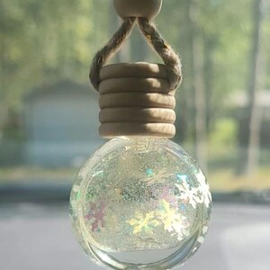Puede incluir: Un ambientador de coche redondo y transparente, con una parte superior de madera y una cuerda de cáñamo. El globo de cristal está lleno de purpurina y confeti con forma de copo de nieve. El fondo está borroso.