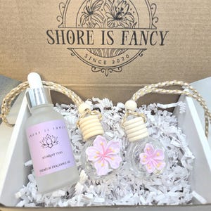 Op de afbeelding: Een geschenkdoos met een matglazen fles Midnight Tides geurolie en twee glazen auto-luchtverfrissers met roze bloemaccenten. De doos is voorzien van het opschrift "Shore is Fancy Since 2020".