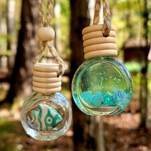 Pode incluir: Dois ambientadores de carro de vidro transparente com tampas de madeira e cordões para pendurar. Um contém um peixe turquesa e branco, o outro tem decorações em forma de concha azul e turquesa. Fundo desfocado.