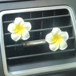 May include: Two white and yellow plumeria flower car air fresheners. The flower-shaped air fresheners are clipped onto the car's air vent. The car vent is black with a silver trim.