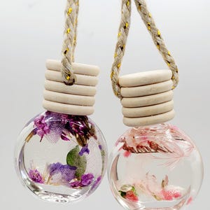 Puede incluir: Dos ambientadores para coche esféricos y transparentes. Uno contiene flores secas moradas y verdes, el otro rosas. Cada uno tiene una tapa de madera y un cordón trenzado dorado y beige para colgar.
