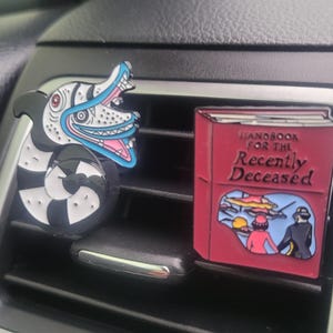 May include: A red car air freshener with the text "Handbook for the Recently Deceased" and a black and white car air freshener with a cartoon monster with a blue mouth.