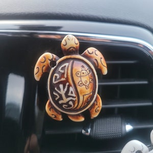 May include: A beige and brown turtle-shaped car air freshener with a decorative design on its shell. The air freshener is hanging from a car's air vent.