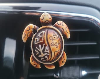 Puede incluir: Un ambientador de coche en forma de tortuga beige y marrón con un diseño decorativo en su caparazón. El ambientador está colgado de una ventilación de aire del coche.