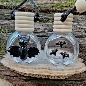 May include: Two clear glass bottles with wooden tops and black cords. One bottle features a black bat, the other has three black bat silhouettes. The bottles are displayed on a piece of wood, suggesting a Halloween or gothic theme.