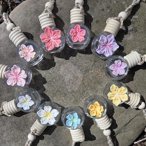 May include: A collection of small, clear glass bottles with colorful flower-shaped decorations inside. Each bottle has a wooden cap and a braided cord for hanging. The flowers are pink, purple, yellow, white, and blue.