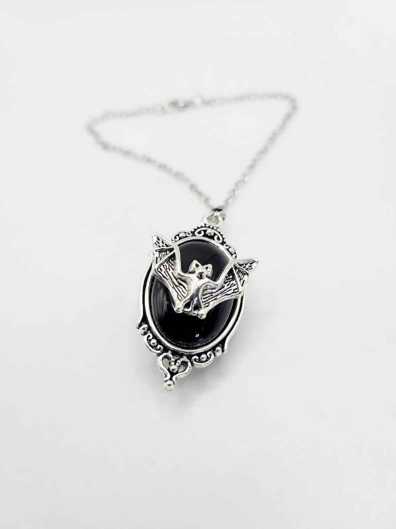 May include: A silver pendant necklace with a black oval cameo featuring a silver bat. The cameo has an ornate silver frame.