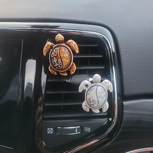 Può includere: Due profumatori per auto decorativi a forma di tartaruga appesi alla bocchetta di ventilazione di un'auto. La tartaruga a sinistra è marrone e beige con un design dettagliato. La tartaruga a destra è argentata con un design simile.