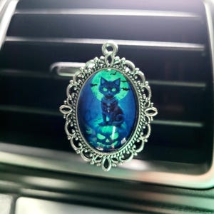 Könnte beinhalten: Ein silberner Auto-Lufterfrischer mit einer schwarzen Katze, die auf einem Kürbis sitzt. Die Katze ist vor einem blauen und grünen Mond silhouettiert.