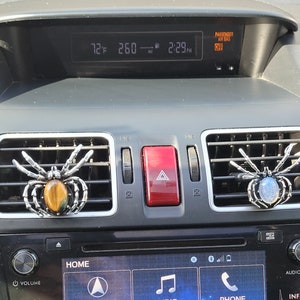 May include: Two silver spider car air fresheners hanging from the air vents of a car. One spider has a tiger eye stone in its abdomen and the other has a blue stone.