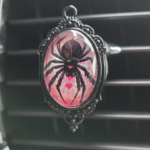 May include: Black spider air freshener with a pink and white web design in a black oval frame with a heart in the centre.