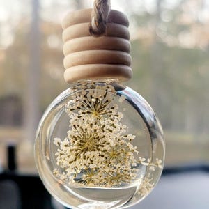 Puede incluir: Ambientador de coche redondo y transparente con flores blancas secas suspendidas en líquido. La parte superior de madera está unida a un cordón trenzado marrón. El fondo está borroso, sugiriendo un entorno exterior.