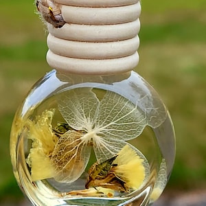 May include: A clear, round glass bottle with dried yellow flowers and a delicate, white, skeletal leaf suspended in a clear liquid. The bottle has a wooden cap and a rope for hanging.