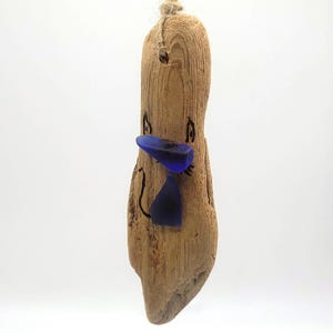 May include: A whimsical driftwood ornament featuring a cartoon-like face. The wooden piece has a natural, light brown color, with a blue glass beak and bow tie. Black lines create the eyes and mouth. A twine loop is attached for hanging.