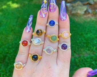 May include: A hand displays a collection of gemstone rings. Each ring features a different colored stone, including red, blue, green, and pink, set in silver or gold wire. The background is a blurred green.