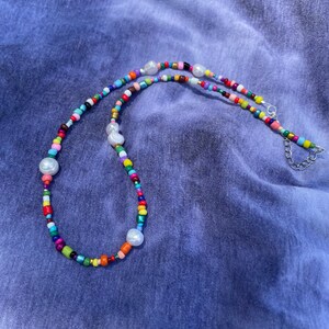 May include: A colorful beaded necklace with a variety of small, round beads in multiple colors, including red, yellow, green, blue, and pink. The necklace also features several large, white, irregularly shaped pearl beads. The necklace is on a blue background.
