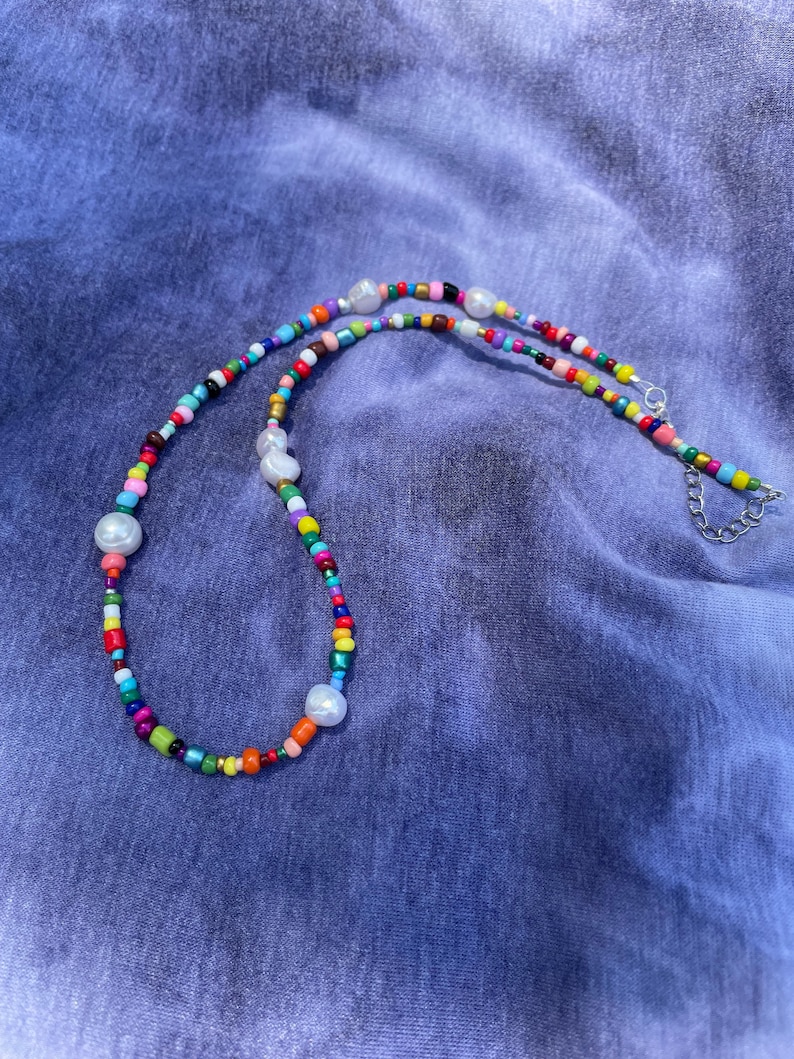 May include: A colorful beaded necklace with a variety of small, round beads in multiple colors, including red, yellow, green, blue, and pink. The necklace also features several large, white, irregularly shaped pearl beads. The necklace is on a blue background.