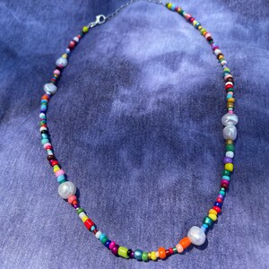 May include: A colorful beaded necklace with a variety of small, round beads in shades of red, orange, yellow, green, blue, and purple. The necklace also features several large, irregular-shaped white pearls and a silver-colored clasp.