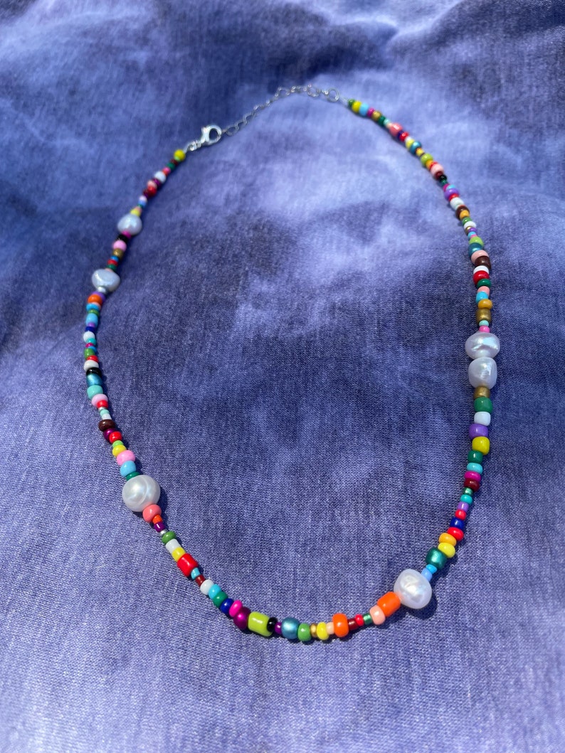 May include: A colorful beaded necklace with a variety of small, round beads in shades of red, orange, yellow, green, blue, and purple. The necklace also features several large, irregular-shaped white pearls and a silver-colored clasp.