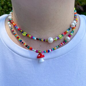 May include: Two beaded necklaces with a colorful array of beads. One necklace features a red and white mushroom pendant. The other necklace has smiley face beads and large white pearls. The necklaces are worn around the neck.