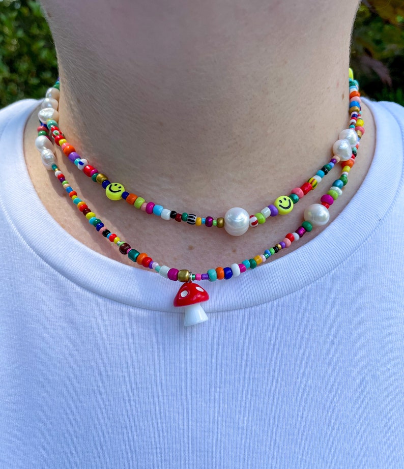 May include: Two beaded necklaces with a colorful array of beads. One necklace features a red and white mushroom pendant. The other necklace has smiley face beads and large white pearls. The necklaces are worn around the neck.