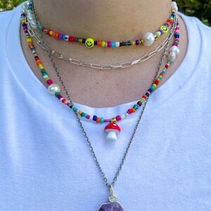 May include: A layered necklace set featuring colorful beaded chokers with smiley faces and pearls, a silver chain, and a pendant with a red mushroom charm. The bottom necklace has a large purple amethyst stone.