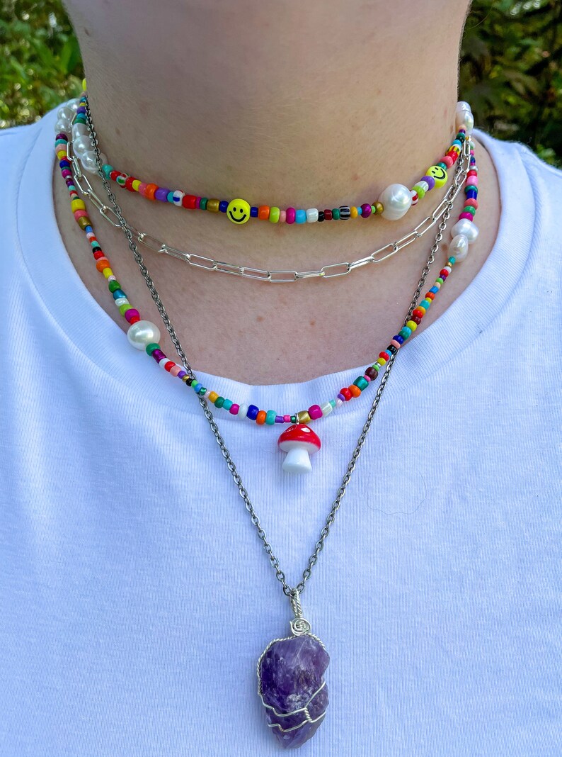 May include: A layered necklace set featuring colorful beaded chokers with smiley faces and pearls, a silver chain, and a pendant with a red mushroom charm. The bottom necklace has a large purple amethyst stone.