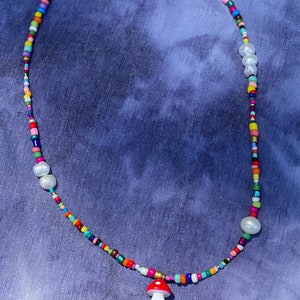 May include: A colorful beaded necklace with a mushroom pendant. The necklace features a variety of small, brightly colored beads, interspersed with larger white pearls. The mushroom pendant is red with white spots and a white base. The necklace has a silver chain.