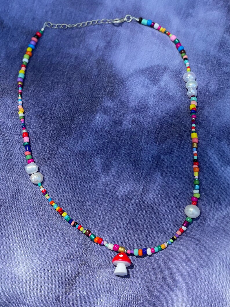 May include: A colorful beaded necklace with a mushroom pendant. The necklace features a variety of small, brightly colored beads, interspersed with larger white pearls. The mushroom pendant is red with white spots and a white base. The necklace has a silver chain.