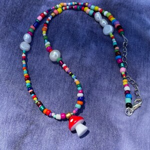 May include: A colorful beaded necklace with a mushroom pendant. The necklace features a variety of small, round beads in multiple colors, interspersed with larger, white pearl-like beads. The mushroom pendant is red with white spots and a white stem.