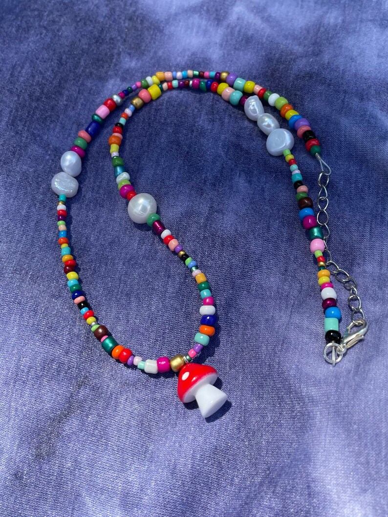 May include: A colorful beaded necklace with a mushroom pendant. The necklace features a variety of small, round beads in multiple colors, interspersed with larger, white pearl-like beads. The mushroom pendant is red with white spots and a white stem.