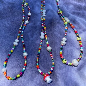 May include: Three colorful beaded necklaces with white pearl accents and silver clasps. One necklace features a red and white mushroom charm, and another has yellow smiley face beads.