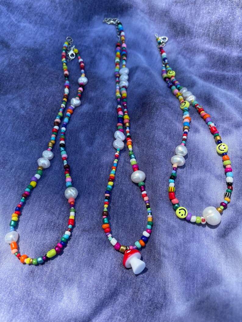 May include: Three colorful beaded necklaces with white pearl accents and silver clasps. One necklace features a red and white mushroom charm, and another has yellow smiley face beads.
