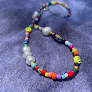 May include: A colorful beaded bracelet with a variety of beads in rainbow colors. The bracelet features large white pearls and yellow smiley face beads. The bracelet is on a blue background.