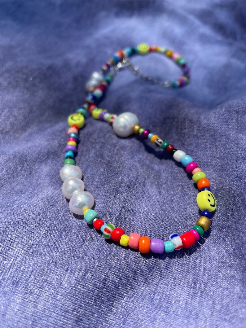 May include: A colorful beaded bracelet with a variety of beads in rainbow colors. The bracelet features large white pearls and yellow smiley face beads. The bracelet is on a blue background.