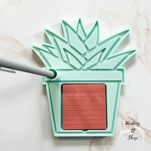 Puede incluir: Un portamemorándum en forma de suculenta verde claro con un bloc de notas rojo. El portamemorándum tiene forma de planta suculenta en maceta. Un bolígrafo gris está insertado en el lateral del portamemorándum. El texto "Making It Shop" está en la esquina inferior derecha.
