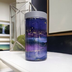 May include: A purple and blue insulated can cooler with a grey lid. The cooler has a galaxy-like design with gold accents.