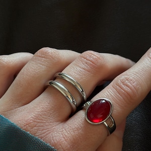 Könnte beinhalten: Zwei silberne Ringe werden an einer Hand gezeigt. Ein Ring hat ein Doppelband-Design, und der andere hat einen großen, ovalen, roten Edelstein, der in einem silbernen Band gefasst ist. Die Ringe werden vor einem dunklen Hintergrund gezeigt.