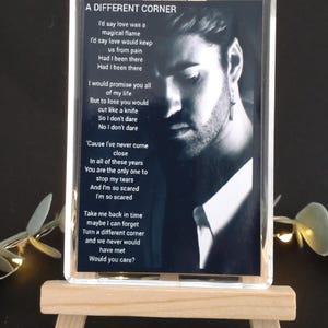 May include: A black and white photograph of a man with lyrics from the song "A Different Corner" by George Michael. The lyrics are printed on a black background and framed in a clear acrylic block.