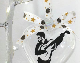 May include: Clear heart-shaped ornament with a black silhouette of a person playing a guitar. The ornament hangs from a white ribbon with gold stars and brown floral accents. A small, glittery heart is attached to the ribbon.