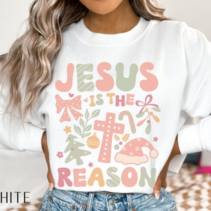 May include: White sweatshirt with a Christmas-themed design featuring the text "Jesus is the reason" in pink, green, and white. The design includes a cross, a bow, a candy cane, a Christmas tree, and a Santa hat.