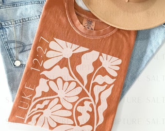May include: A rust-colored t-shirt with a white floral design and the text "LUKE 12:27".