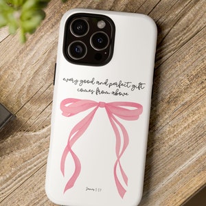 May include: White phone case featuring a pink watercolor bow design and the text "every good and perfect gift comes from above." The case is on a wooden surface, with a camera and plant in the background.