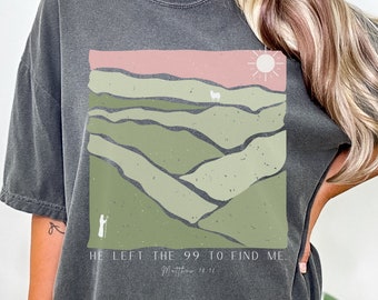 May include: A charcoal grey oversized t-shirt with a graphic print of a green landscape with a pink sunset and a white sun. The text "He left the 99 to find me. Matthew 18:11" is printed below the graphic.