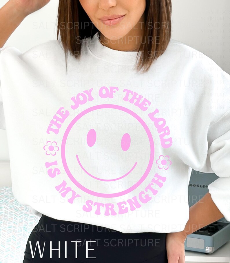 May include: White sweatshirt with a pink smiley face design. The design includes the text "THE JOY OF THE LORD IS MY STRENGTH" in a circular pattern around the smiley face. The word "WHITE" is at the bottom.