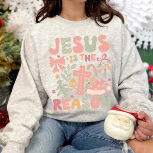 May include: A gray sweatshirt with a Christmas-themed design featuring the text "Jesus is the reason" in pink and green letters. The design also includes a cross, a bow, candy canes, and a Santa hat.