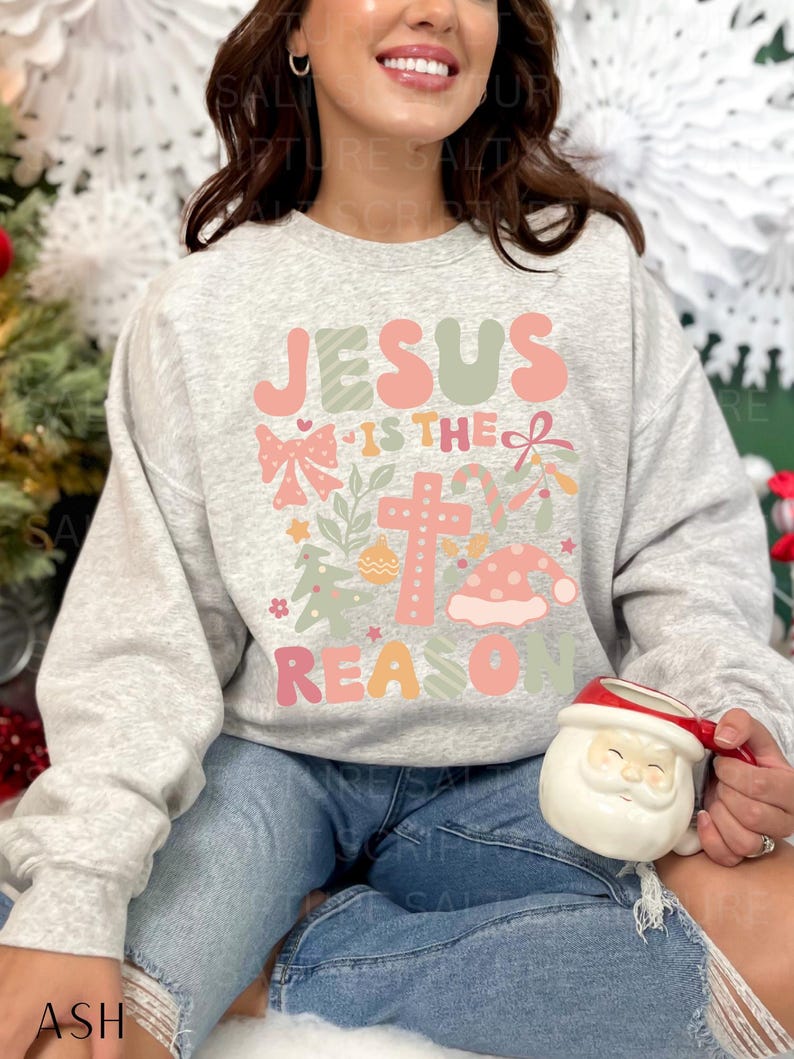 May include: A gray sweatshirt with a Christmas-themed design featuring the text "Jesus is the reason" in pink and green letters. The design also includes a cross, a bow, candy canes, and a Santa hat.
