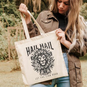 May include: A beige tote bag with black text and a graphic of a lion's head with a crown. The text on the bag reads "Hail Hail Lion of Judah Let the Lion Roar".