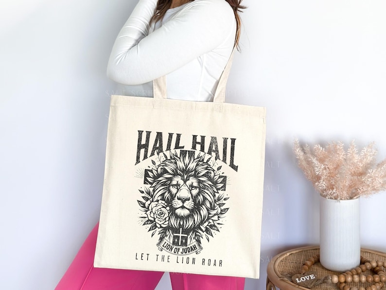 May include: A white tote bag with a black and white graphic of a lion's head with the text "Hail Hail Let the Lion Roar" and "Lion of Judah" printed on it.